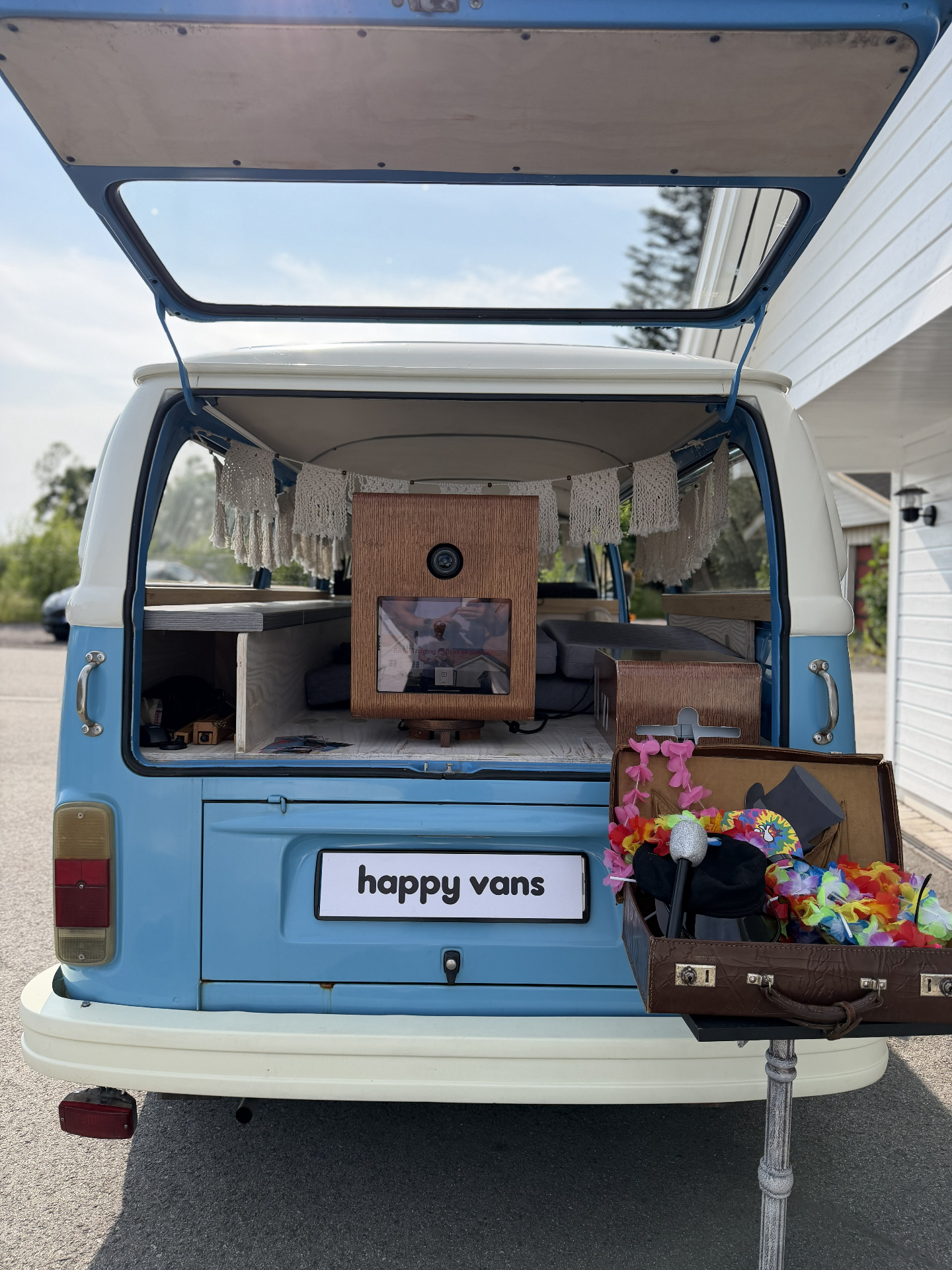 Bakluken på lyseblå VW-buss åpen med HappySmiles photobooth og koffert full av hatter, blomsterkranser og briller ved siden av