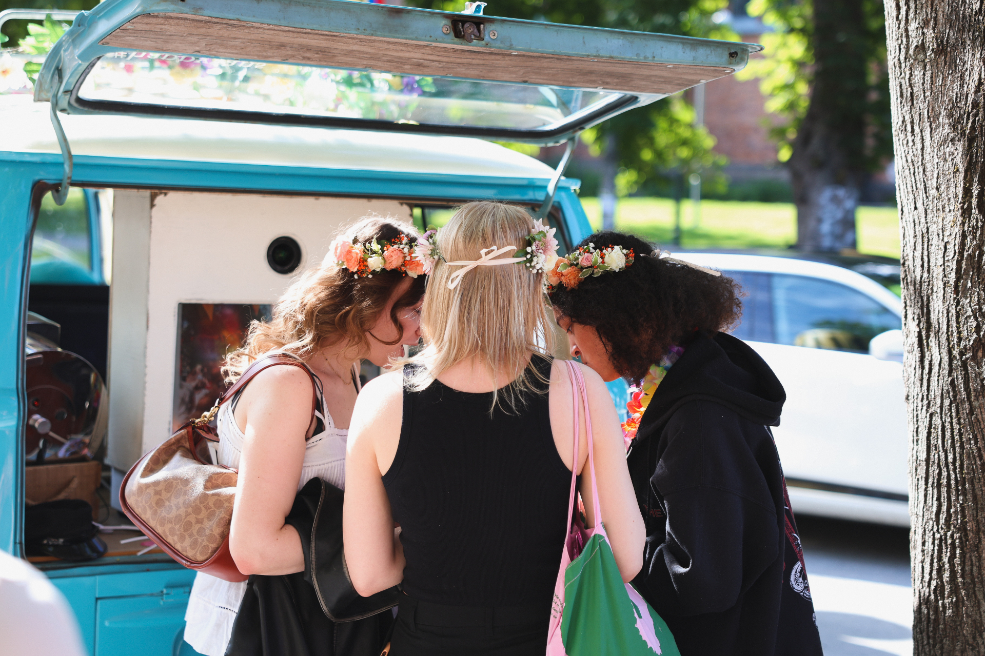 Tre venninner med blomsterkranser studerer ferske utskrifter ved HappySmiles VW-buss-booth