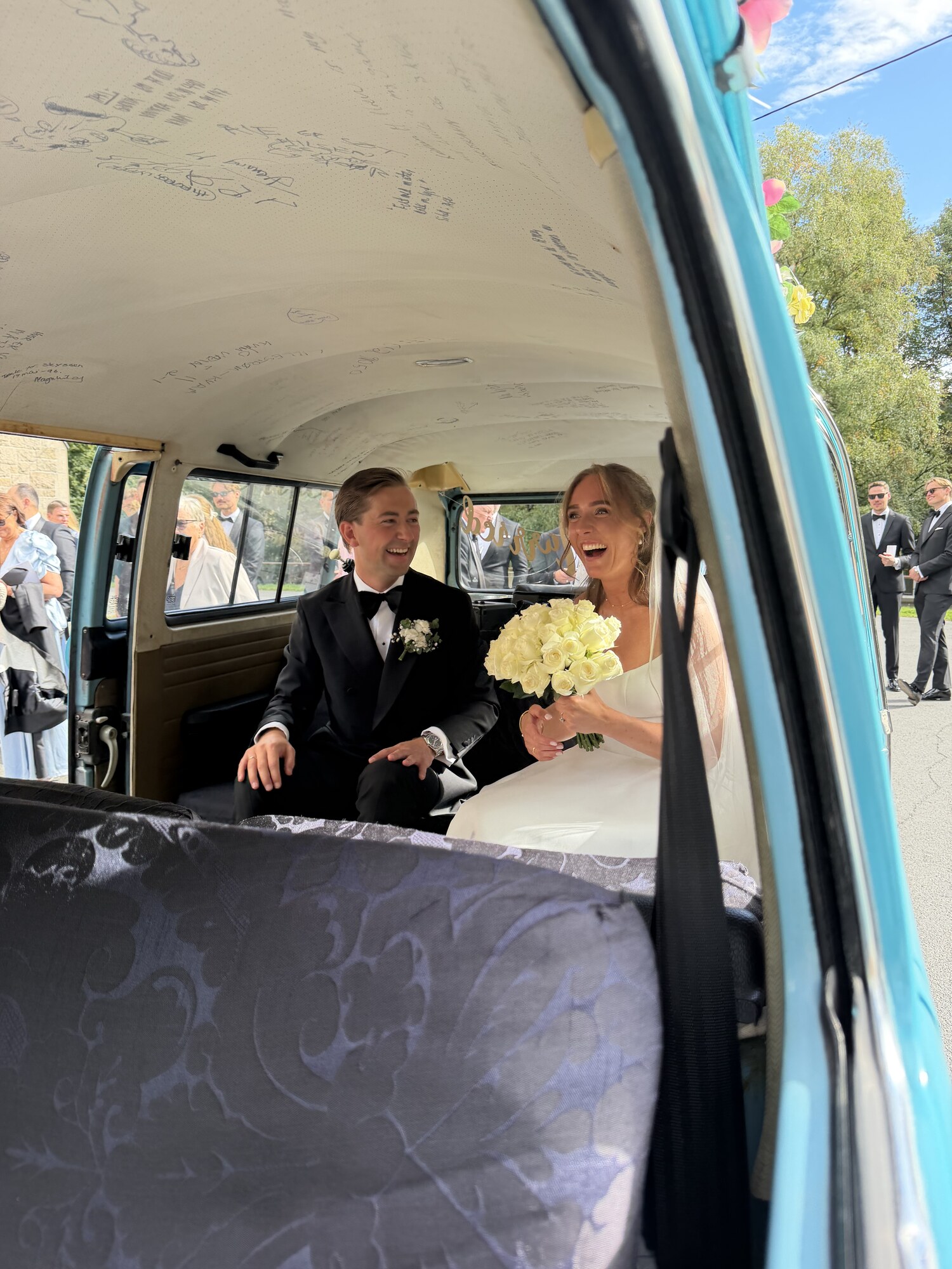 Brudeparet Anette og Roger sitter smilende bak i den turkise VW-bussen med brudebukett og Just Married-pynt