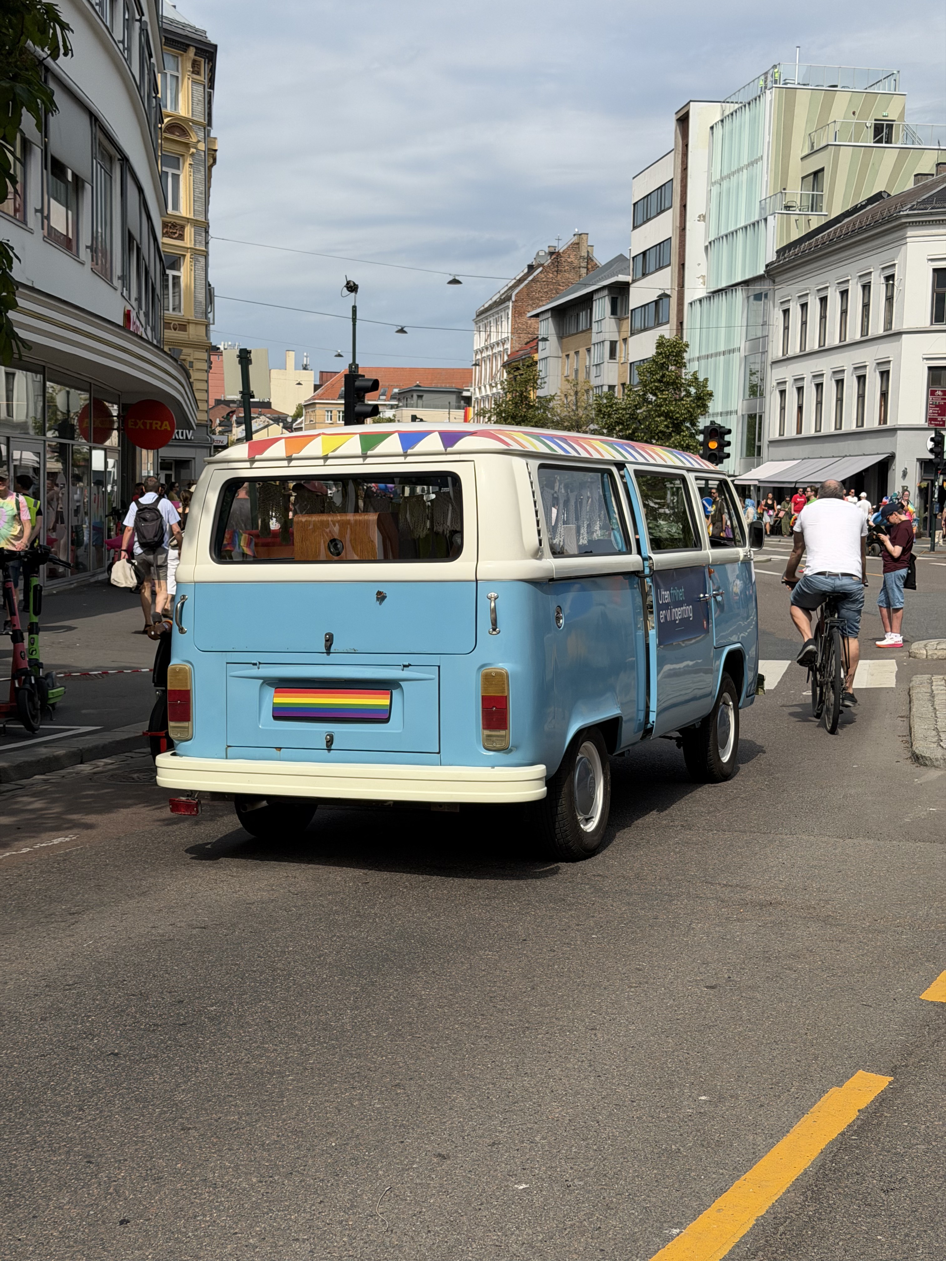 Lyseblå VW-buss bakfra med regnbuevimpler på taket og regnbueflagg på bakluken, midt i en Oslo-gate under Pride-paraden