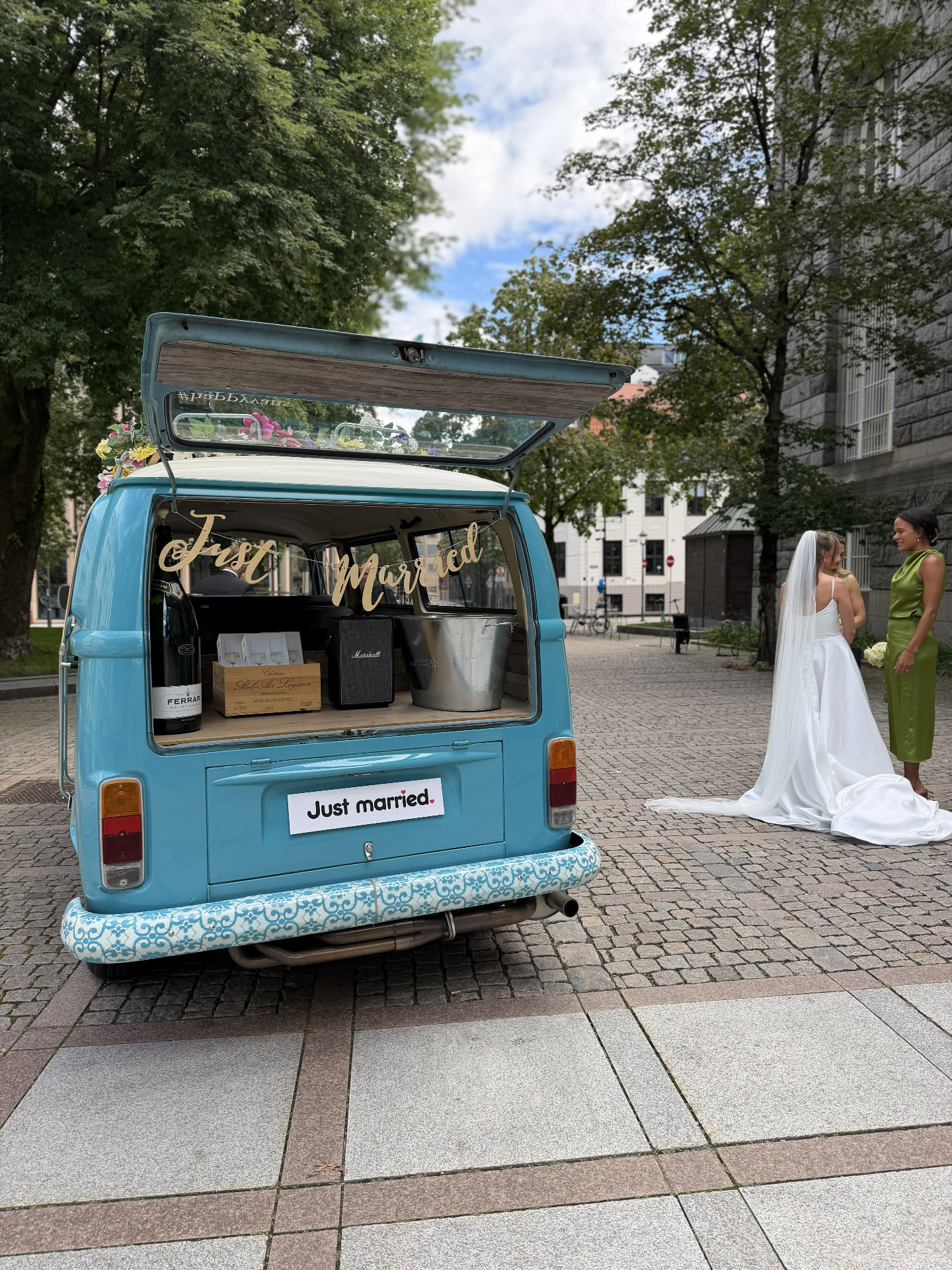 Bruden i hvit kjole ved siden av den turkise VW-bussen med åpen bakluke, champagne og Just Married-skilt på Bankplassen