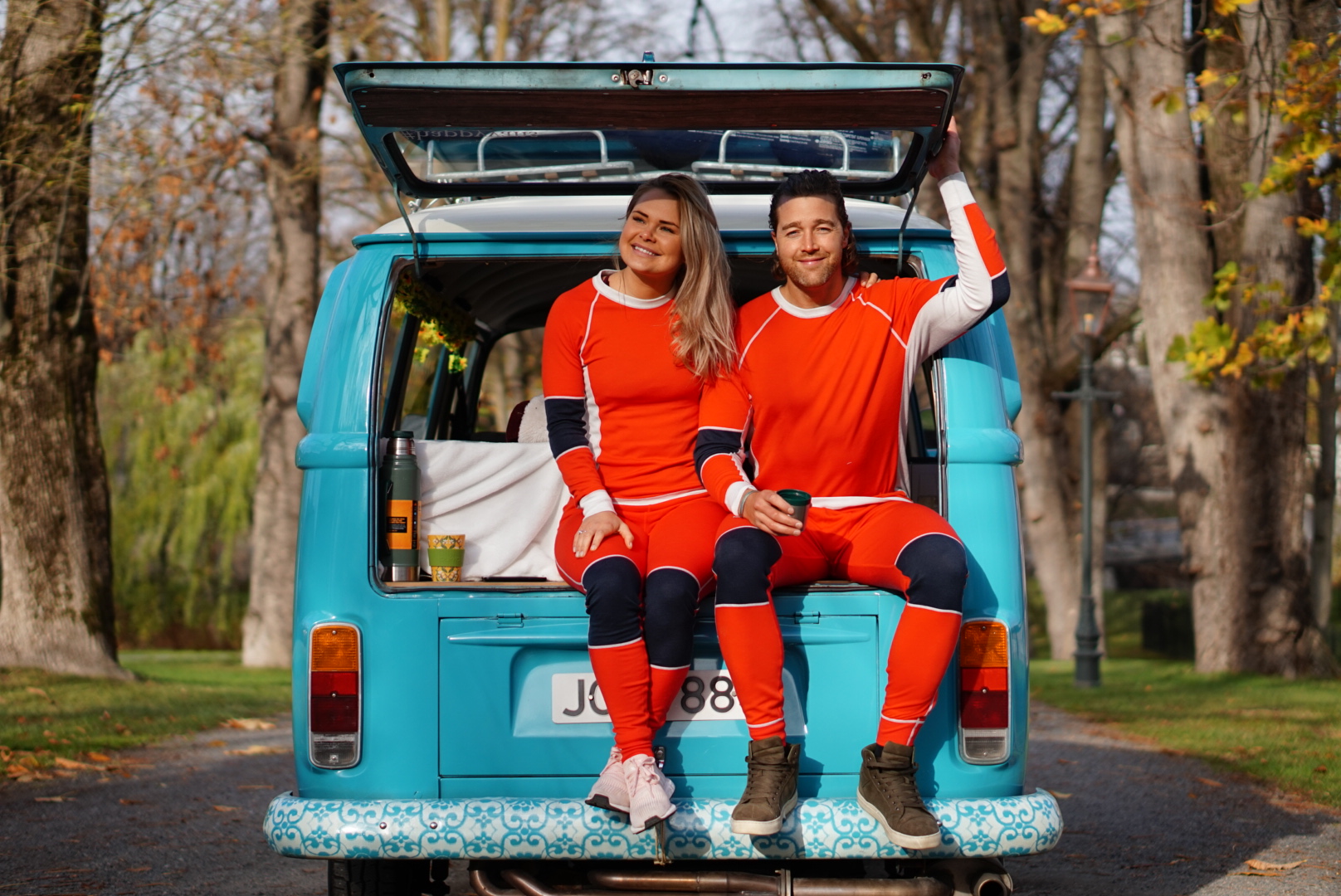 To smilende teammedlemmer i oransje antrekk sitter bak i en turkis VW-buss på en allé om høsten