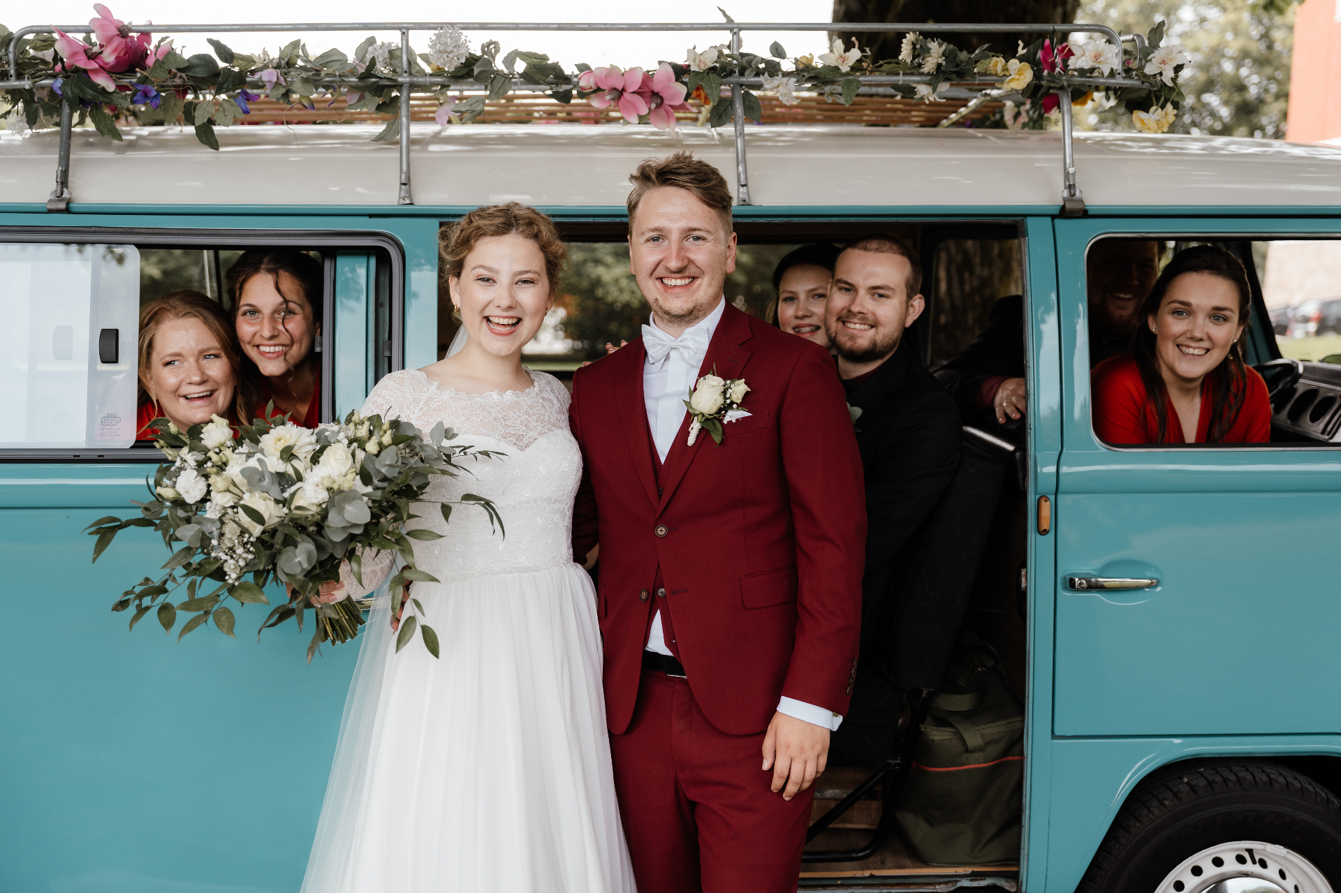 Smilende brudepar foran turkis VW-buss med blomsterpyntet takgrind og hele følget i vinduene
